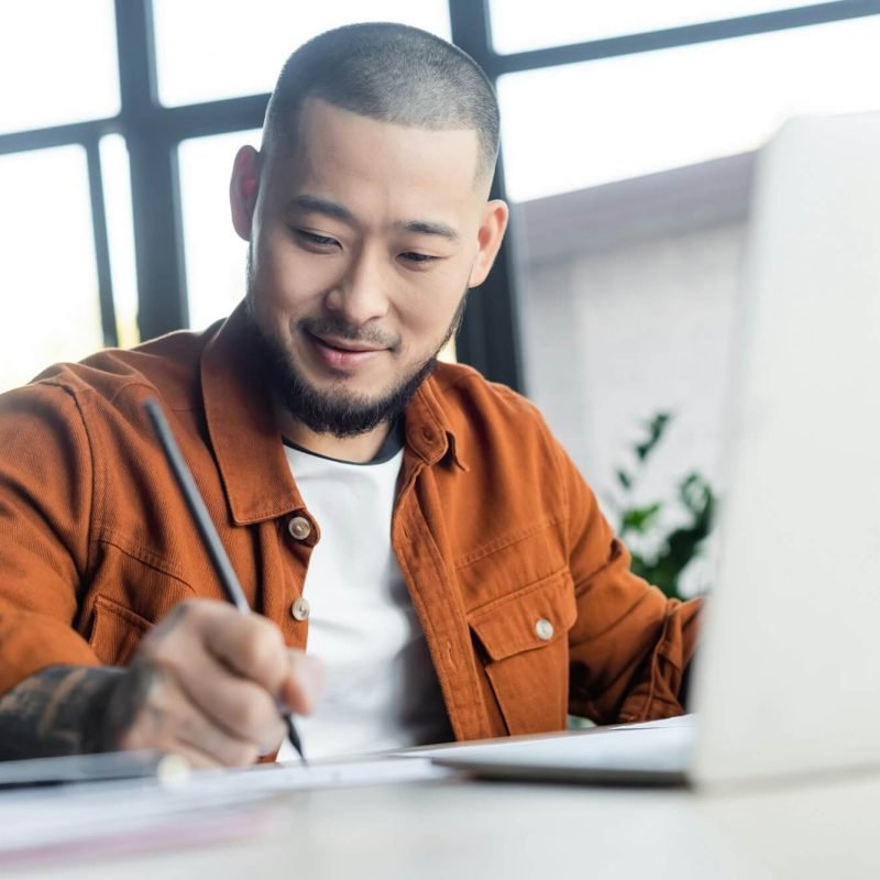 smiling-asian-designer-drawing-near-blurred-laptop-in-architectural-bureau.jpg